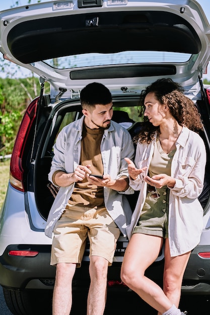 Pareja joven tranquila en ropa casual sentada en el maletero del coche y discutiendo la ruta de su viaje mientras camina el fin de semana de verano