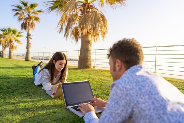 Pareja joven tumbado en la hierba en el trabajo inteligente con el portátil uno frente al otro al atardecer o al amanecer.