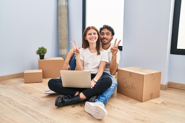 Foto pareja joven usando laptop en casa nueva sonriendo con cara feliz guiñando un ojo a la cámara haciendo el signo de la victoria con los dedos número dos