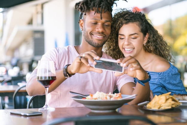 Pareja joven usando un teléfono móvil y tomando fotos de la comida mientras almuerzan juntos en un restaurante.