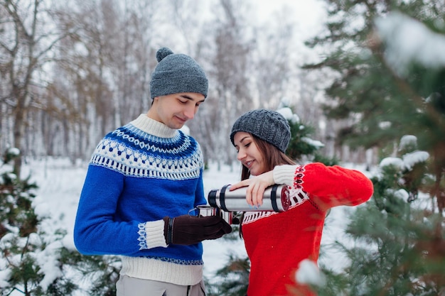 Pareja joven vertiendo té caliente de un termo en el bosque de invierno Gente feliz relajándose al aire libre durante las vacaciones