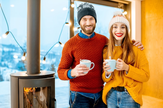 Pareja joven vestida con suéteres brillantes y sombreros de pie junto con bebidas calientes cerca de la chimenea en la casa moderna durante el invierno