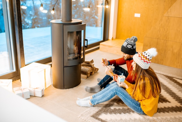 Pareja joven vestida con suéteres y sombreros brillantes sentados juntos cerca de la chimenea en la casa moderna durante el invierno