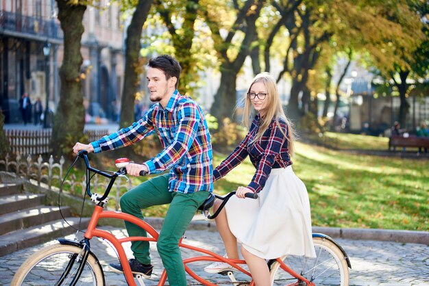 Pareja joven viajero feliz activo, hombre guapo con barba y mujer bonita rubia en bicicleta tándem en un día soleado en la escalera pedregosa y el antiguo edificio de fondo.