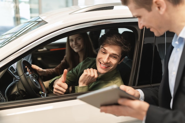 Una pareja joven visitando un salón de autos sentado dentro de un automóvil hablando con un asistente de ventas mirando