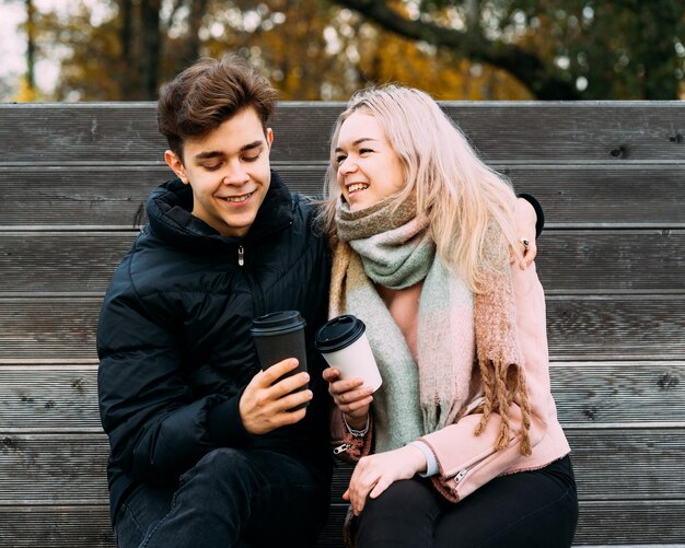 Foto una pareja de jóvenes alegres sosteniendo tazas de café desechables mientras están sentados en un banco en el parque