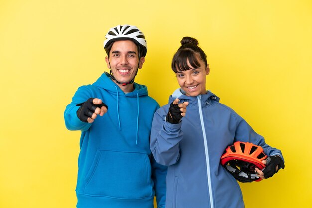 Una pareja de jóvenes ciclistas aislada de fondo amarillo te señala con una expresión de confianza