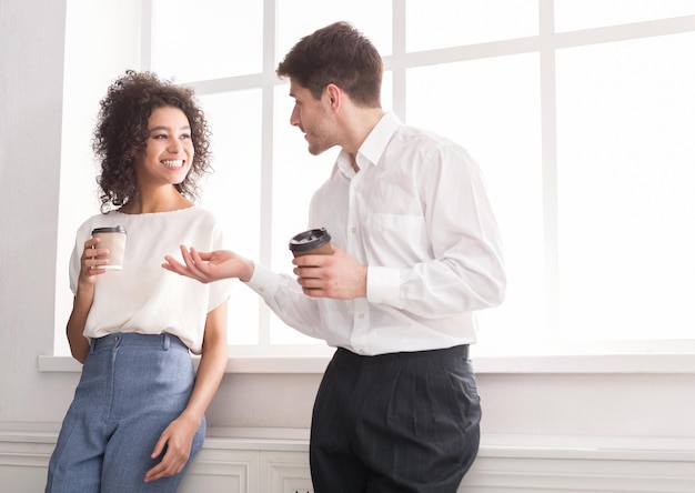 Pareja de jóvenes colegas de pie junto a la ventana y tomando café