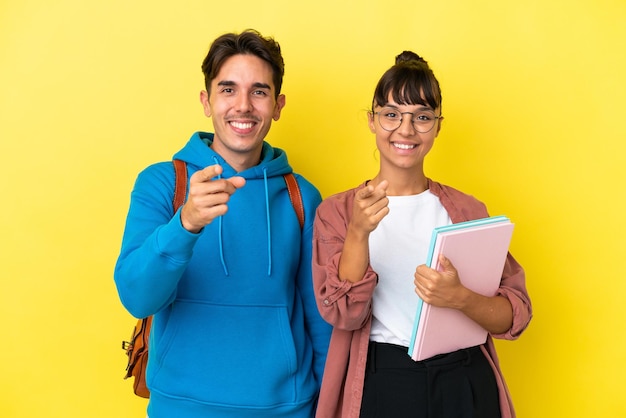 Una pareja de jóvenes estudiantes aislada de fondo amarillo te señala con una expresión de confianza