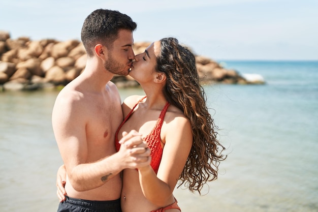 Pareja de jóvenes hispanos turistas vistiendo traje de baño bailando en la playa