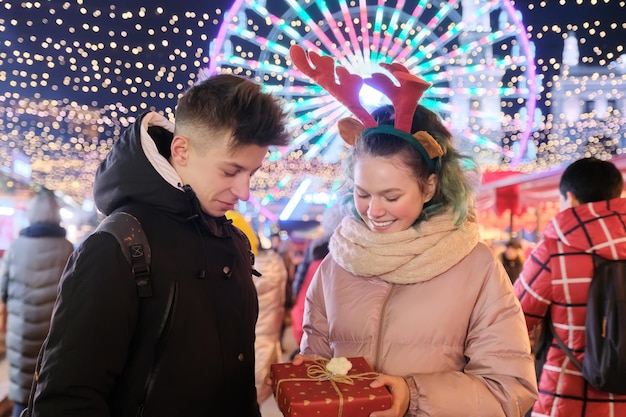 Pareja de jóvenes en el mercado navideño. Feliz día de fiesta niño y niña dando caja de regalo. Adolescentes hablando riendo, luces brillantes de fondo de guirnaldas de la ciudad de noche, noria