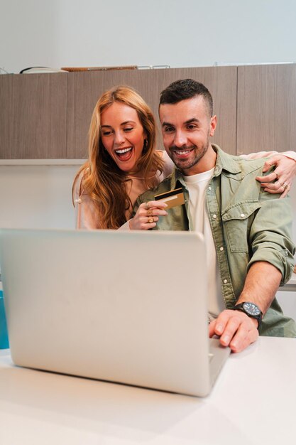 Una pareja de jóvenes verticales sonriendo mientras reservan sus vacaciones de ensueño juntos usando una computadora portátil y crédito