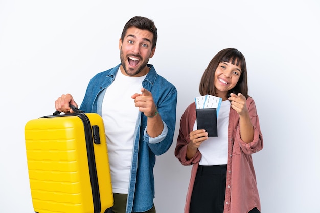 Una pareja de jóvenes viajeros que sostienen una maleta y un pasaporte aislados de fondo blanco te señalan con una expresión de confianza