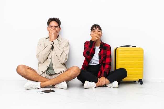 Una pareja de jóvenes viajeros de raza mixta sentada en el suelo aislada de fondo blanco sonriendo mucho mientras cubría la boca