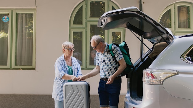 Pareja jubilada cargando equipaje de viaje en el maletero del coche mientras se va de vacaciones de verano en el aniversario del matrimonio. Turistas mayores preparándose para un viaje de campo mientras colocan equipaje y carritos dentro del vehículo