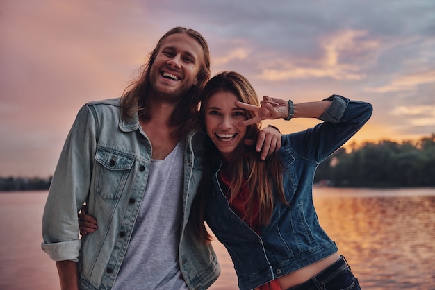 Pareja juguetona. Feliz pareja joven en ropa casual sonriendo y mirando a la cámara mientras está de pie cerca del lago