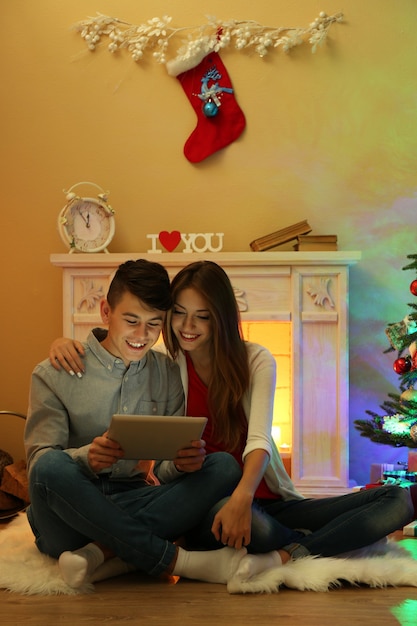 Pareja junto a la chimenea en casa decorada de Navidad con tableta