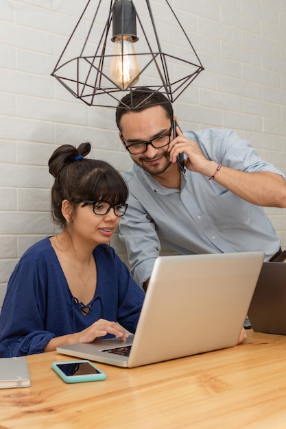 Pareja latina de empresarios trabajando en casa. hombre hablando por teléfono mientras la mujer trabaja en una laptop en una larga mesa de madera