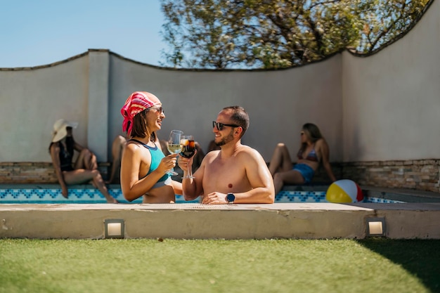 Una pareja latina sonriente brinda dentro de una piscina rodeada de amigos