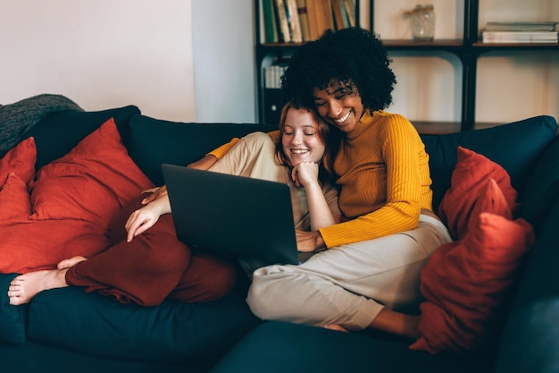 Pareja lesbiana relajada femenina multiétnica en el sofá viendo la computadora portátil y la vinculación