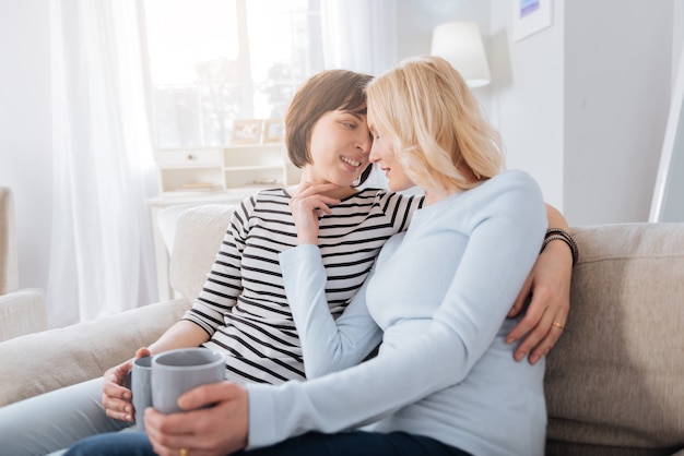 Pareja de lesbianas. Agradable agradable pareja de lesbianas sentada en el sofá y tomando té mientras descansan juntos