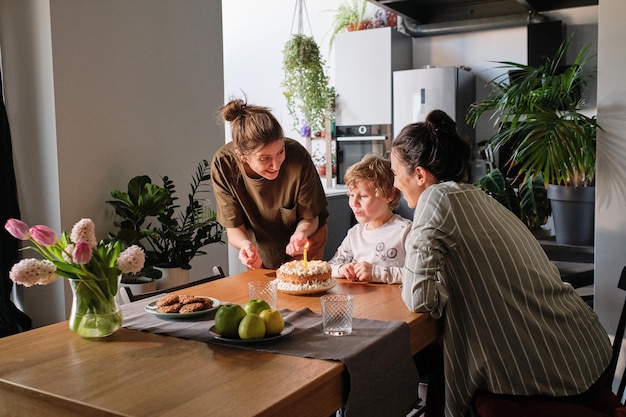 Pareja de lesbianas dando pastel de cumpleaños con velas a su pequeño hijo en la mesa de la cocina