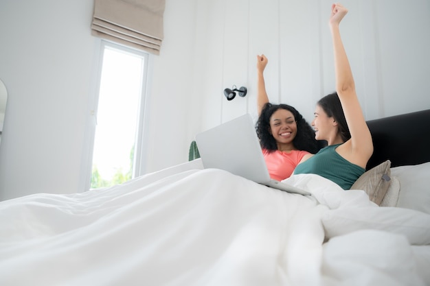 Una pareja de lesbianas felices usando una computadora portátil en la cama en casa. Una pareja de las lesbianas multiétnicas usando una laptop.