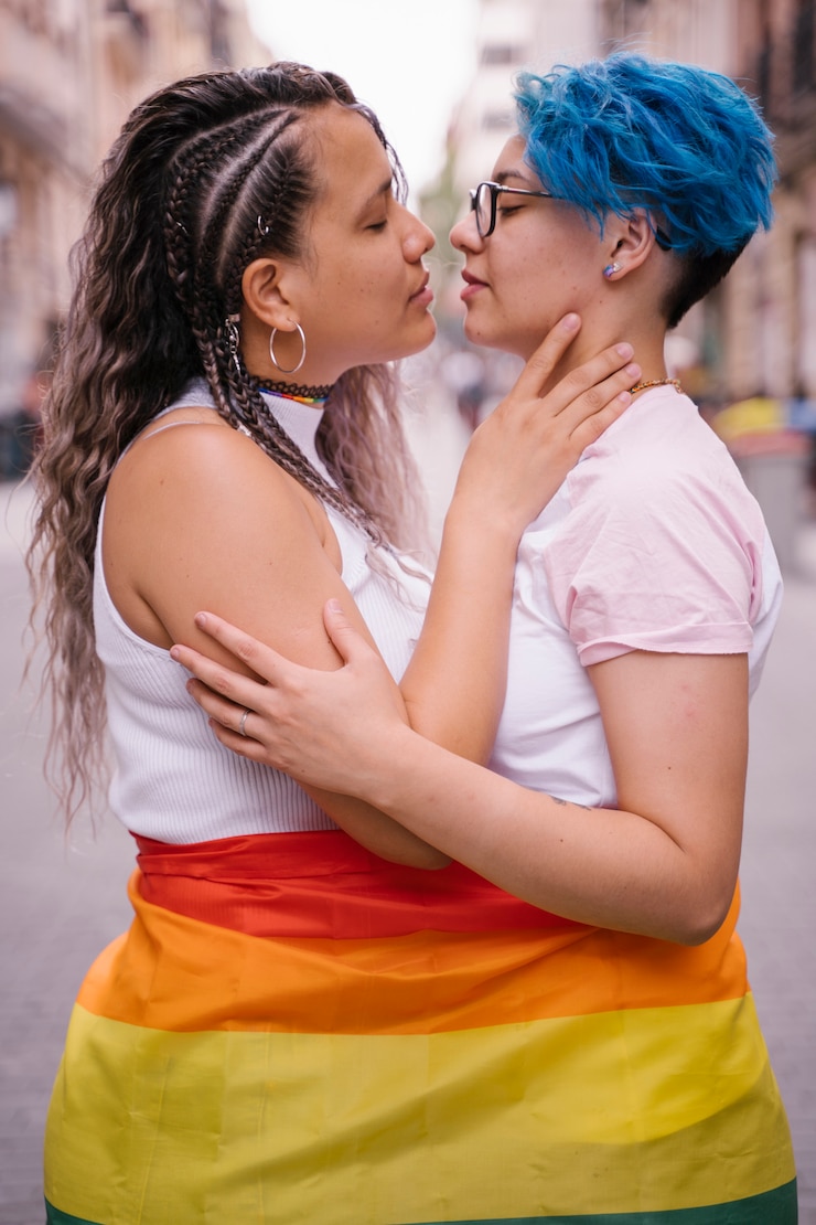 Foto pareja de lesbianas vistiendo con orgullo una bandera lgbt y besándose en la calle.