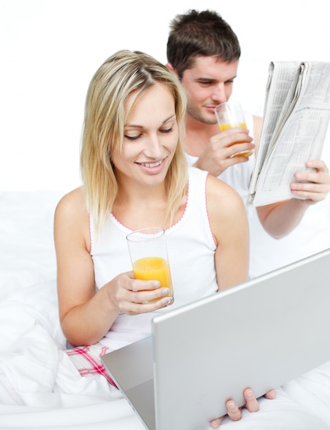 Pareja leyendo un periódico y usando una computadora portátil en la cama