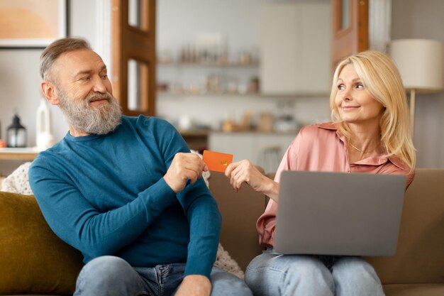 Pareja madura de adictos a las compras de compras con computadora portátil y tarjeta de crédito interior