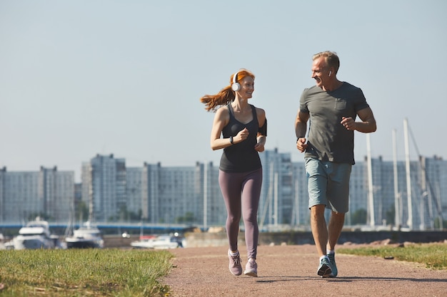 Pareja madura deportiva en auriculares sonriendo el uno al otro mientras disfruta de trotar juntos en la ciudad