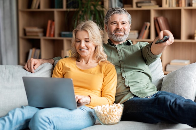 Pareja madura descansando en el sofá en casa viendo la televisión y usando la computadora portátil