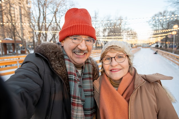 Pareja madura sonriente haciendo retrato selfie mientras patina en pista de patinaje durante las vacaciones de invierno