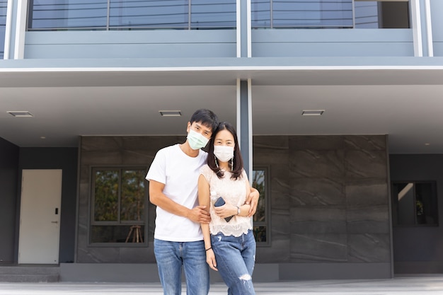 Pareja con máscara protectora de pie y abrazándose frente a su nueva casa.