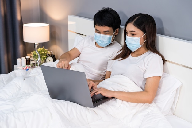 Pareja con máscaras médicas que usan una computadora portátil en la cama durante el coronavirus pandémico en cuarentena se quedan en casa.