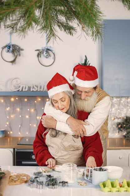 Pareja mayor en delantales y gorros rojos navideños horneando galletas en la cocina