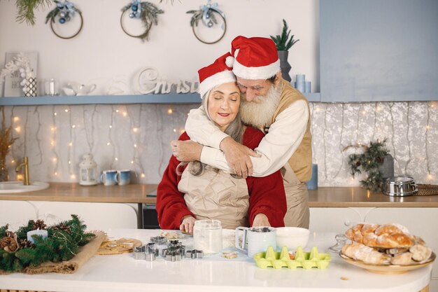 Pareja mayor en delantales y gorros rojos navideños horneando galletas en la cocina