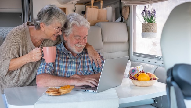 Pareja mayor despreocupada en vacaciones de viaje sentada dentro de una autocaravana disfrutando del desayuno usando lapt