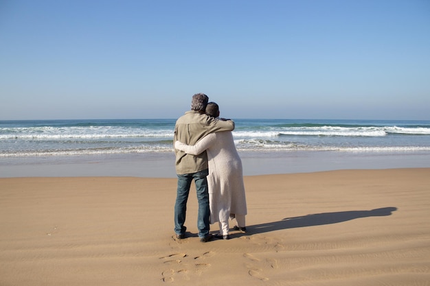 Una pareja mayor disfruta de una hermosa vista del paisaje marino mientras se para en la arena mojada y se abraza. Cónyuges de mediana edad que pasan vacaciones juntos en la playa. Jubilación, amor, concepto de viaje.