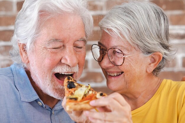 Pareja mayor divirtiéndose juntos comiendo pizza en casa fondo de pared de ladrillo