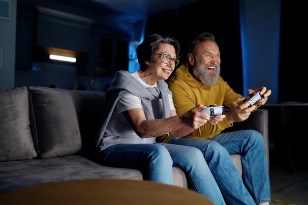 Foto pareja mayor jugando videojuegos juntos en casa esposa y marido ancianos sentados en el sofá sosteniendo joysticks y sintiéndose emocionados por el tiempo libre el fin de semana