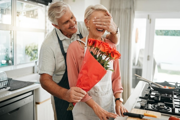 Pareja mayor que cubre los ojos y las flores sorprende cuando el hombre le da a la esposa un ramo de rosas en un cumpleaños de aniversario o en el día de San Valentín en la cocina Feliz anciano y mujer siendo románticos en la casa de Australia