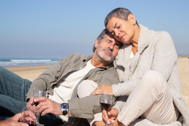 Una pareja mayor relajada disfrutando de un picnic a la orilla del mar en un día soleado. Hombre y mujer de cabello gris sosteniendo copas de vino, apoyándose el uno en el otro y descansando con los ojos cerrados. concepto de ocio