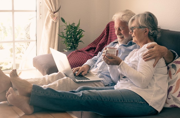 Una pareja mayor relajada tumbada en un sofá en casa usando una laptop Dos ancianos jubilados disfrutando de tiempo libre y dispositivos tecnológicos mujer sosteniendo en la mano una taza de café o té