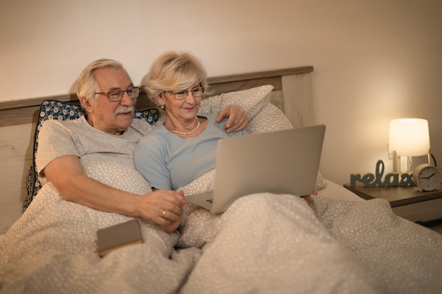 Una pareja mayor sonriente relajándose en el dormitorio y navegando por Internet en una laptop