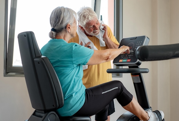 Una pareja de mayores confiada juntos en el gimnasio una mujer haciendo bicicleta de ejercicio y mostrando el resultado en exhibición