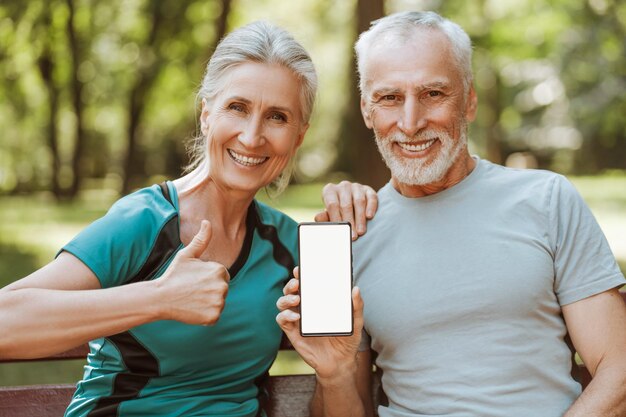 Una pareja de mayores feliz mostrando un teléfono inteligente con una pantalla de maqueta sentada en un banco en el espacio de copia del parque