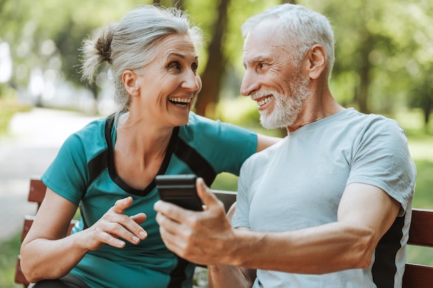 Una pareja de mayores feliz usando un teléfono inteligente sentada en un banco en el parque ordenando la compra de redes sociales