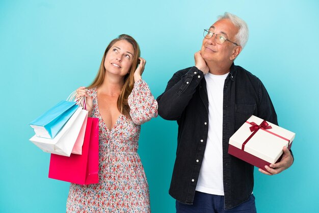 Pareja de mediana edad con bolsa de compras y regalo aislado sobre fondo azul pensando en una idea mientras se rasca la cabeza