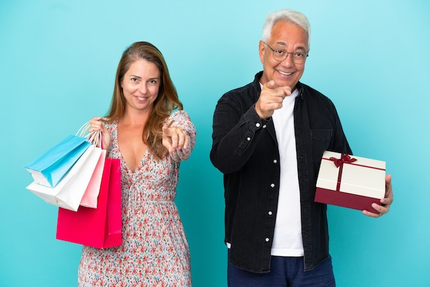 Pareja de mediana edad con bolsa de compras y regalo aislado sobre fondo azul le señala con el dedo con una expresión de confianza